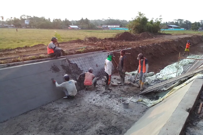 Pekerja konstruksi memplester dinding lereng saluran irigasi tersier trapesium dengan adukan semen dan tulangan wiremesh di persawahan desa